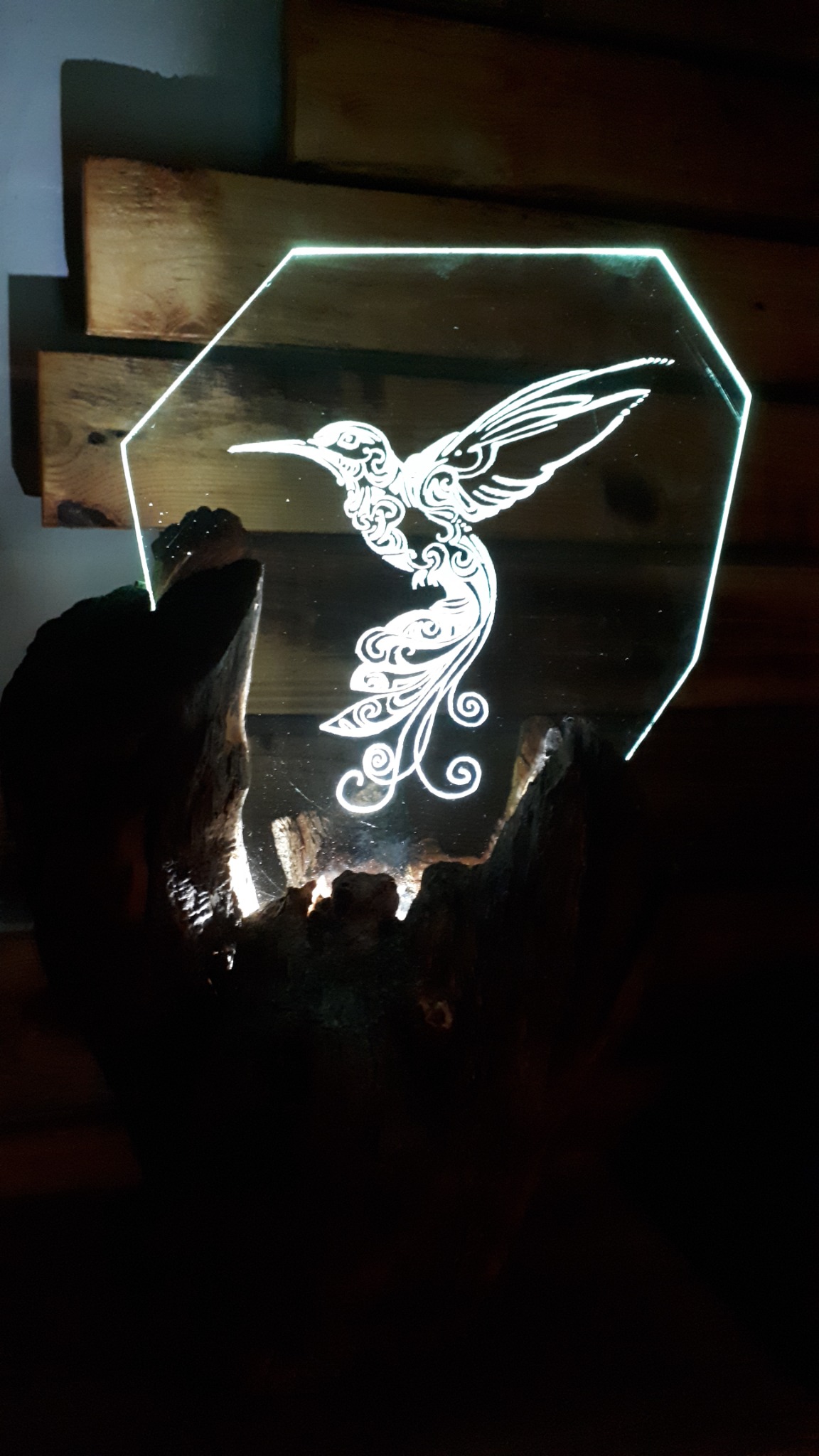 Podświetlany, dekoracyjny panel z pleksi w kształcie ośmiokąta z wygrawerowanym kolibrem, umieszczony w podstawie z surowego drewna.