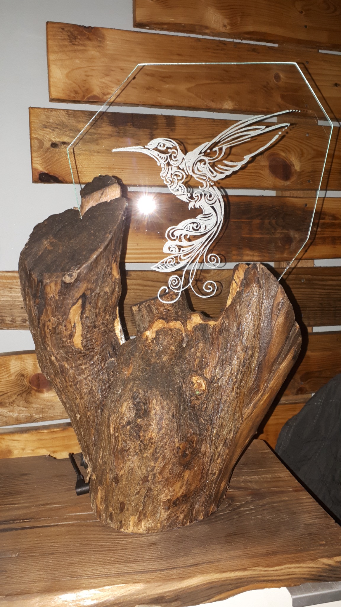 Oryginalna lampka nocna: pień z transparentną płytą z wygrawerowanym kolibrem na tle drewnianej ściany.