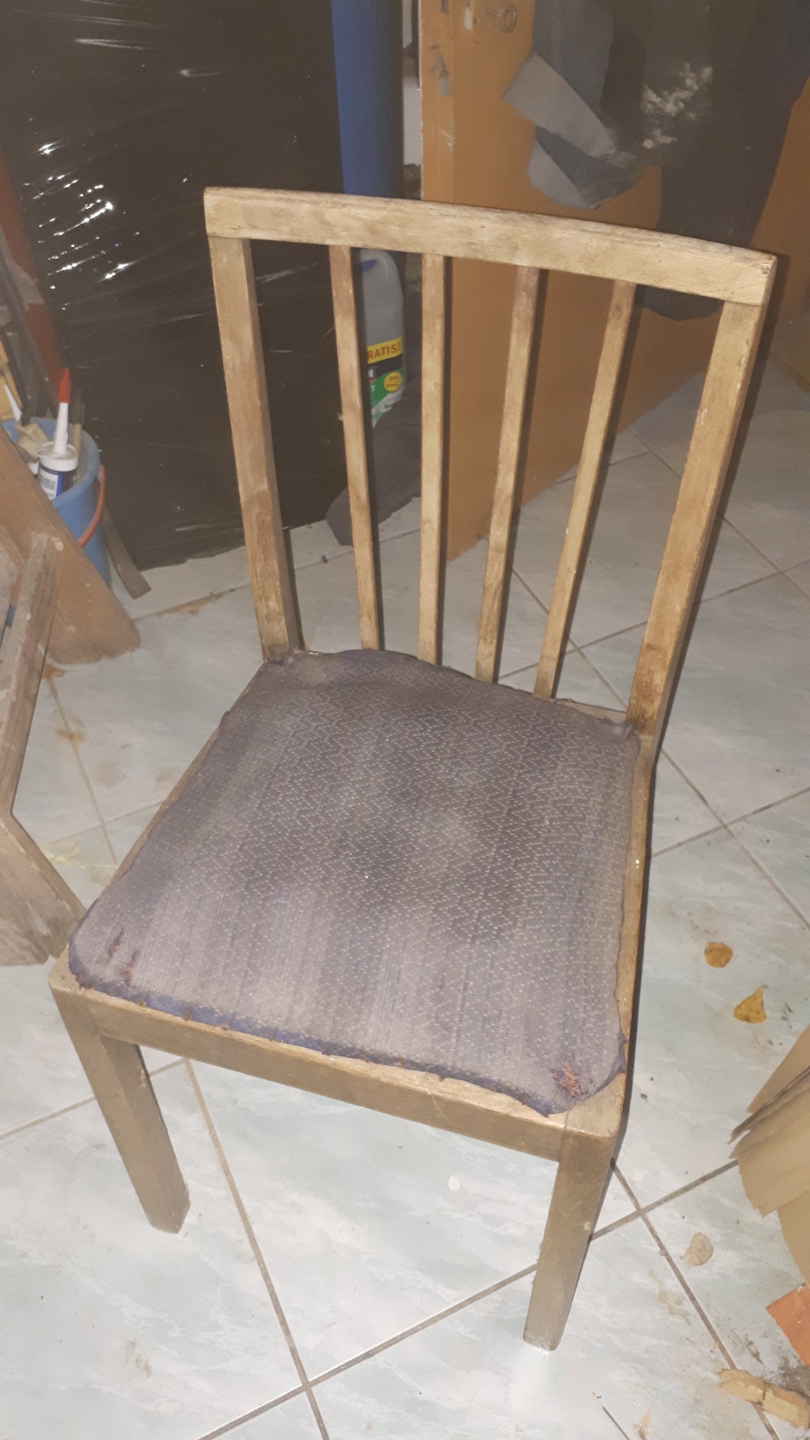 Stare, drewniane krzesło z tapicerowanym siedziskiem w odcieniach szarości, widoczne ślady użytkowania i zabrudzenia na siedzisku, tło stanowią materiały budowlane i narzędzia.