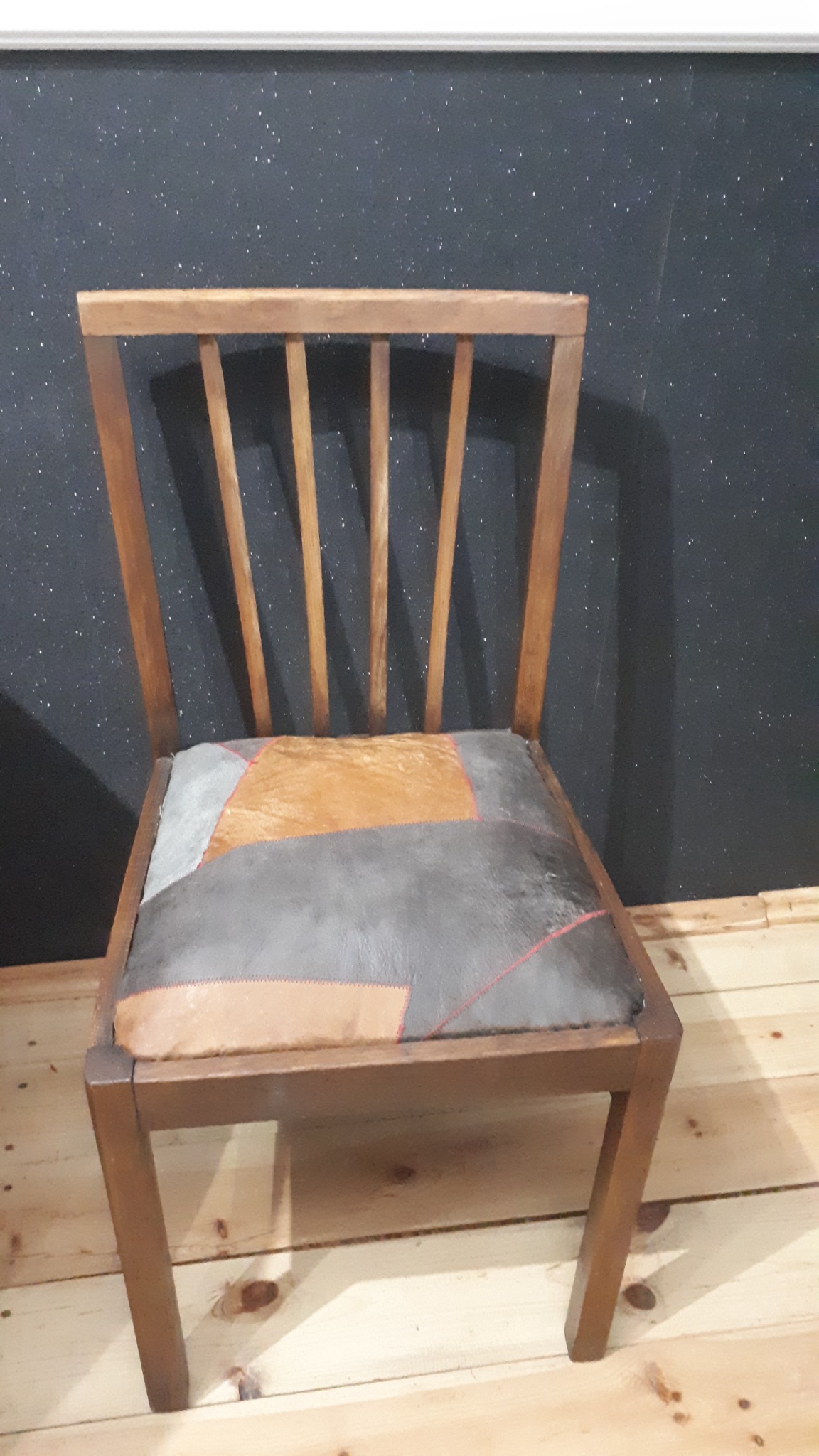 Drewniane krzesło z patchworkowym siedziskiem z brązowej i szarej skóry, stojące na jasnej drewnianej podłodze na tle ciemnej ściany z drobnymi wzorami.