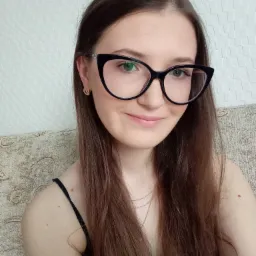 Portret uśmiechniętej brunetki w czarnych okularach o kocim kształcie, siedzącej na beżowej kanapie z wzorem, na tle białej ściany z teksturą.