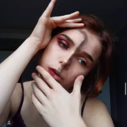 Młoda kobieta z połową twarzy pomalowaną ciemną farbą i intensywnym makijażem oka w odcieniach czerwieni, pozująca z dłońmi przy twarzy.