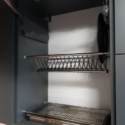 Wnętrze szafki kuchennej z szarymi frontami, wyposażonej w metalowy ociekacz na naczynia dwupoziomowy, z widocznymi mechanizmami podnoszenia frontu.