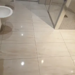 Beżowe, prostokątne płytki podłogowe w łazience, widoczny fragment toalety, bidetu i kabiny prysznicowej z przezroczystymi drzwiami.