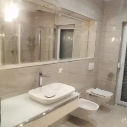 Nowoczesna łazienka w beżowych odcieniach z podwieszaną umywalką na białym blacie, bidetem i toaletą, widoczna duża lustrzana ściana i kabina prysznicowa w odbiciu.