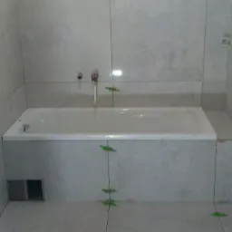 Wanna obudowana płytkami w trakcie montażu, widoczne krzyżyki dystansowe i sznurki poziomujące, ściany również wyłożone płytkami w odcieniach szarości.