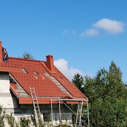 Radekglazewski26 - Częściowo zdjęte czerwone dachówki na domu z białego pustaka. Dwóch dekarzy pracuje przy kominie, a obok stoją drabiny. W tle błękitne niebo i drzewa.