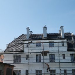 Radekglazewski26 - Budynek w trakcie ocieplania, widoczne rusztowania i pracownik na nich. Dach pokryty czarną dachówką. Białe kominy. Błękitne niebo w tle.
