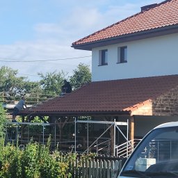 Radekglazewski26 - Montaż brązowej dachówki na drewnianej konstrukcji altany. Dwóch mężczyzn pracuje na rusztowaniu. W tle dom z czerwonym dachem i białą elewacją.