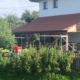 Radekglazewski26 - Montaż brązowej dachówki na drewnianej konstrukcji z rusztowaniem. Widoczny fragment budynku i ogrodzenie z zielenią. Prace dekarskie w toku, dekarz na dachu.