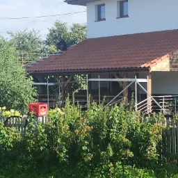 Montaż brązowej dachówki na drewnianej konstrukcji z rusztowaniem. Widoczny fragment budynku i ogrodzenie z zielenią. Prace dekarskie w toku, dekarz na dachu.