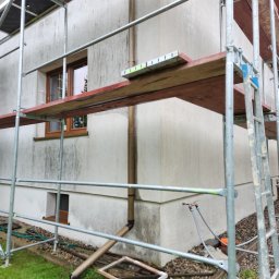 Radekglazewski26 - Elewacja budynku w trakcie czyszczenia pod rusztowaniem. Widoczne zabrudzenia na ścianie, rynna i fragment trawnika. Ujęcie z poziomu gruntu.