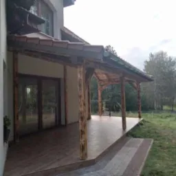 Zadaszony taras z drewnianymi słupami i fragmentem dachu z brązową dachówką, przylegający do jasnego budynku. Widok na zielony ogród.