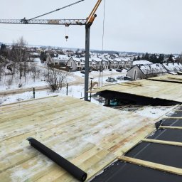 Radekglazewski26 - Budowa dachu w zimowej scenerii. Widoczna drewniana konstrukcja, czarna membrana dachowa i żuraw budowlany w tle osiedla domów pokrytych śniegiem.
