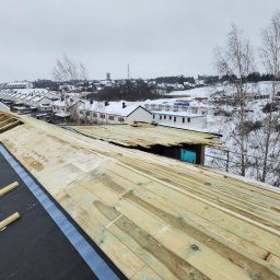Radekglazewski26 - W trakcie budowy dachu: widok na drewnianą konstrukcję więźby dachowej pokrytą czarną membraną, na tle zimowego krajobrazu z zabudowaniami i drzewami.