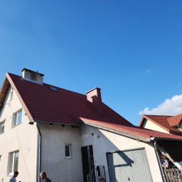 Radekglazewski26 - Dom jednorodzinny z czerwonym dachem z blachodachówki w słoneczny dzień. Widoczny komin i okno dachowe. Częściowo widoczni dwaj mężczyźni.