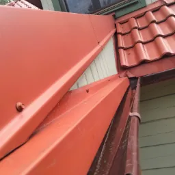 Czerwona blacha okapowa połączona z dachówką ceramiczną, widoczny fragment rynny spustowej i okno dachowe