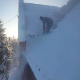Pracownik w czerwonej kurtce usuwa śnieg z dachu budynku za pomocą piły łańcuchowej, widoczne rusztowanie i drzewa w tle.