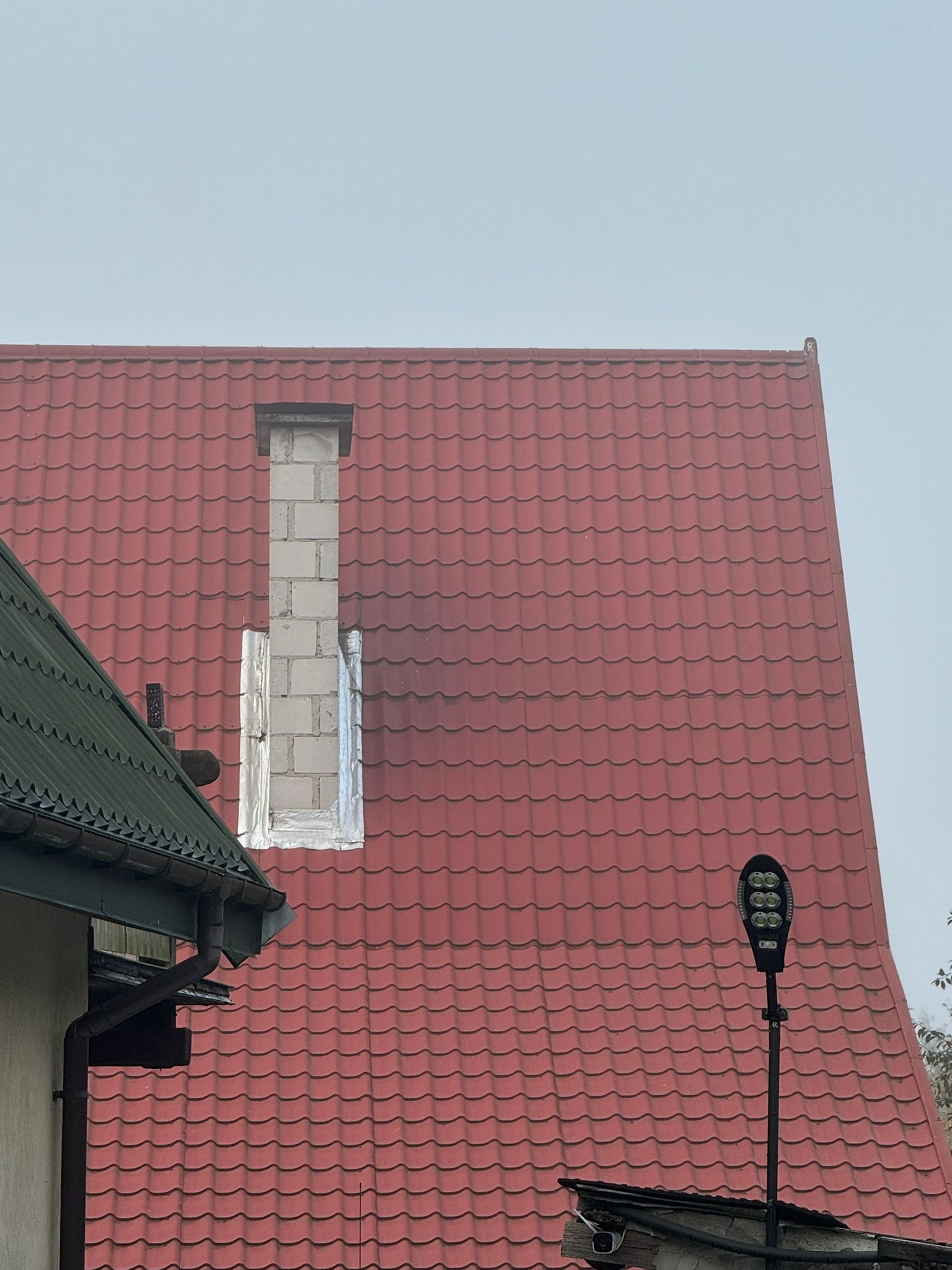 Czerwony dach z kominem w trakcie budowy, zabezpieczony folią. Widoczny fragment rynny i lampa uliczna. Ujęcie z dołu.