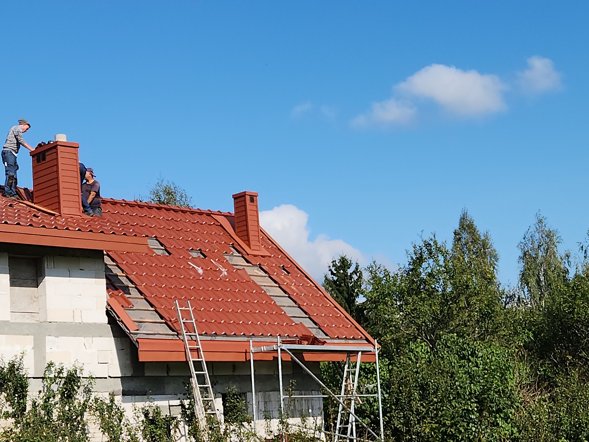 Częściowo zdjęte czerwone dachówki na domu z białego pustaka. Dwóch dekarzy pracuje przy kominie, a obok stoją drabiny. W tle błękitne niebo i drzewa.