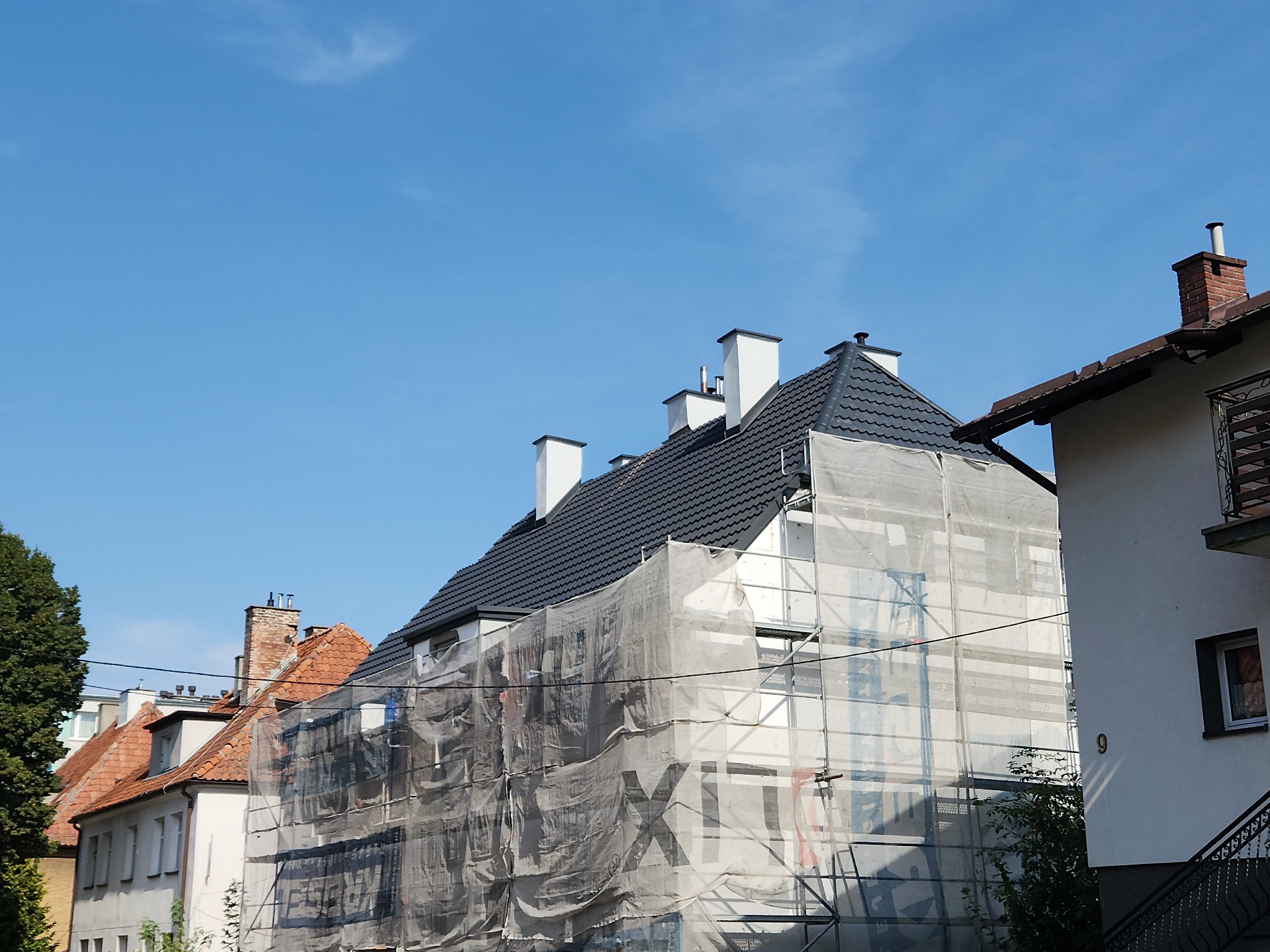 Dom w rusztowaniach, osłonięty siatką, z nowym, ciemnym dachem. Widoczne kominy. Sąsiednie budynki z dachami z czerwonej dachówki w tle, błękitne niebo.