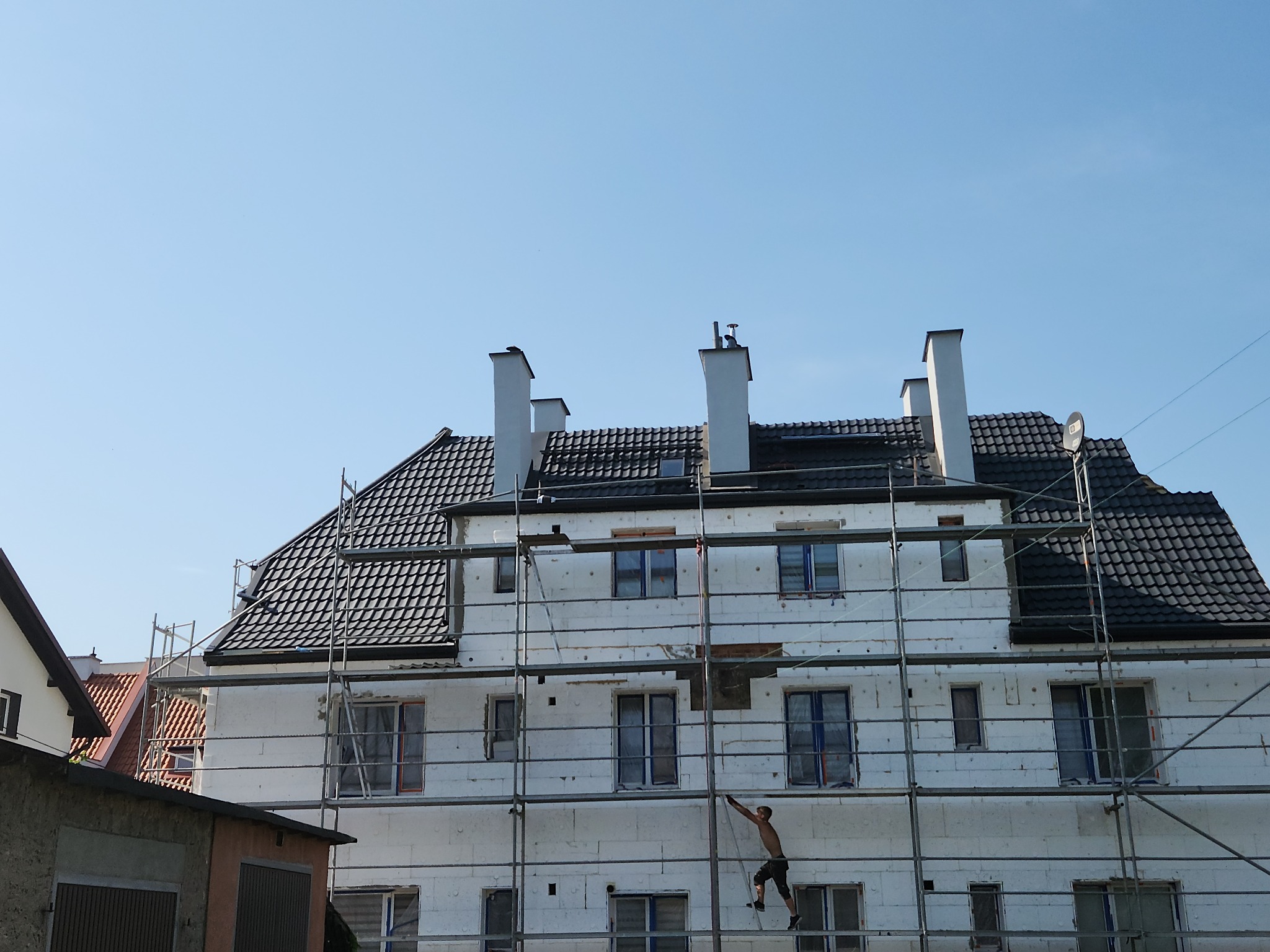 Budynek w trakcie ocieplania, widoczne rusztowania i pracownik na nich. Dach pokryty czarną dachówką. Białe kominy. Błękitne niebo w tle.