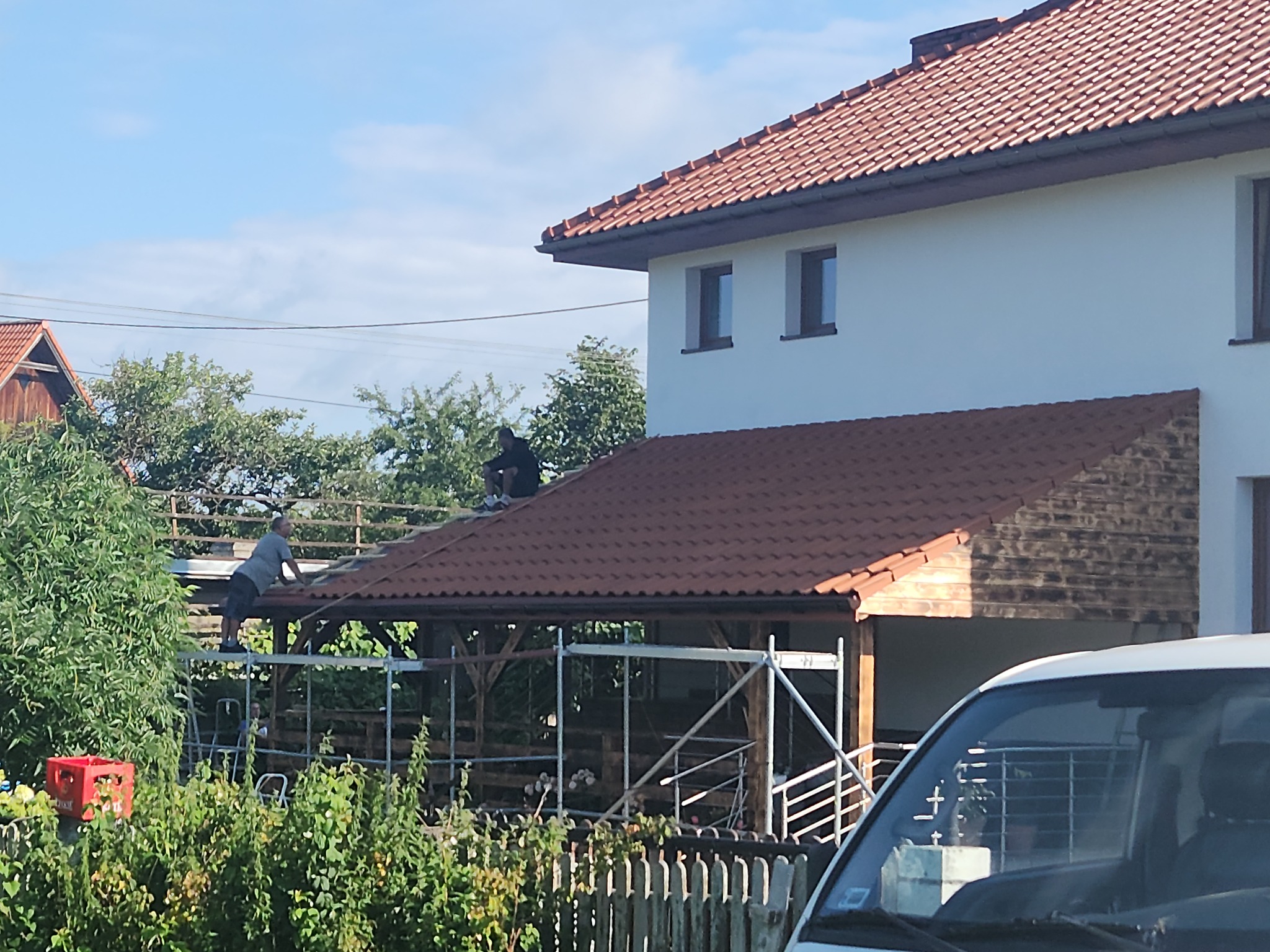 Montaż brązowej dachówki na drewnianej konstrukcji altany. Dwóch mężczyzn pracuje na rusztowaniu. W tle dom z czerwonym dachem i białą elewacją.