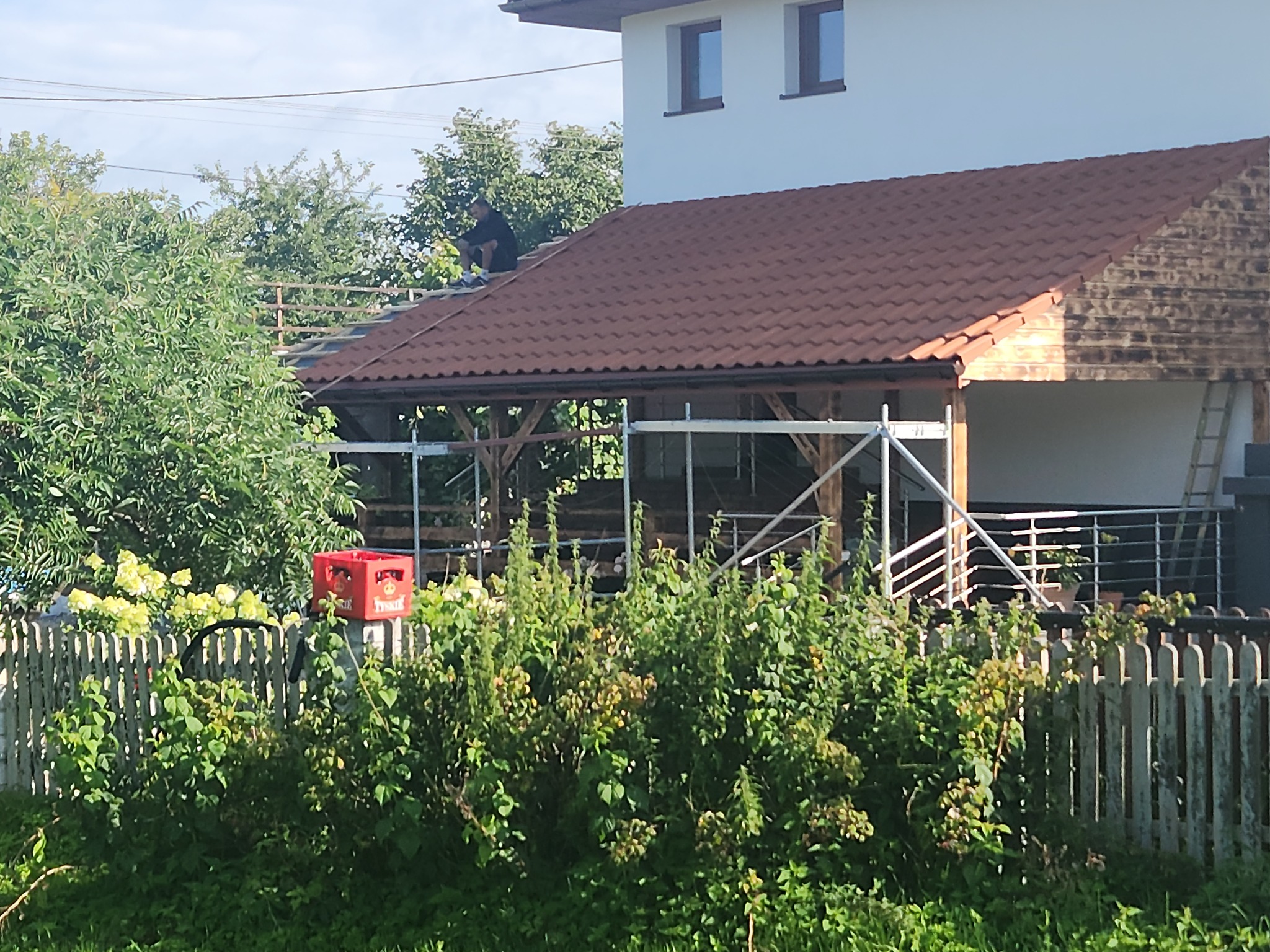 Montaż brązowej dachówki na drewnianej konstrukcji z rusztowaniem. Widoczny fragment budynku i ogrodzenie z zielenią. Prace dekarskie w toku, dekarz na dachu.