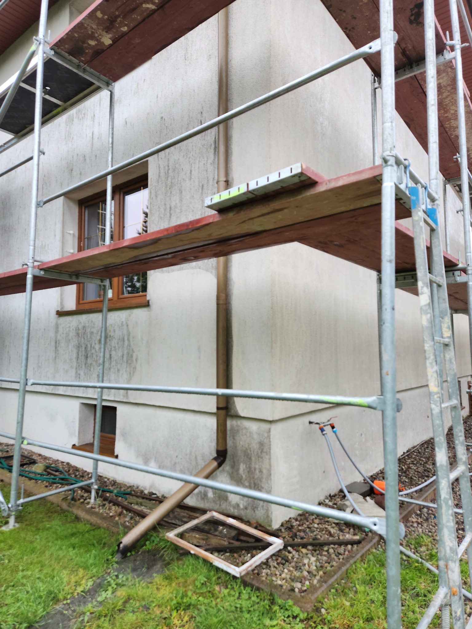 Elewacja budynku w trakcie czyszczenia pod rusztowaniem. Widoczne zabrudzenia na ścianie, rynna i fragment trawnika. Ujęcie z poziomu gruntu.