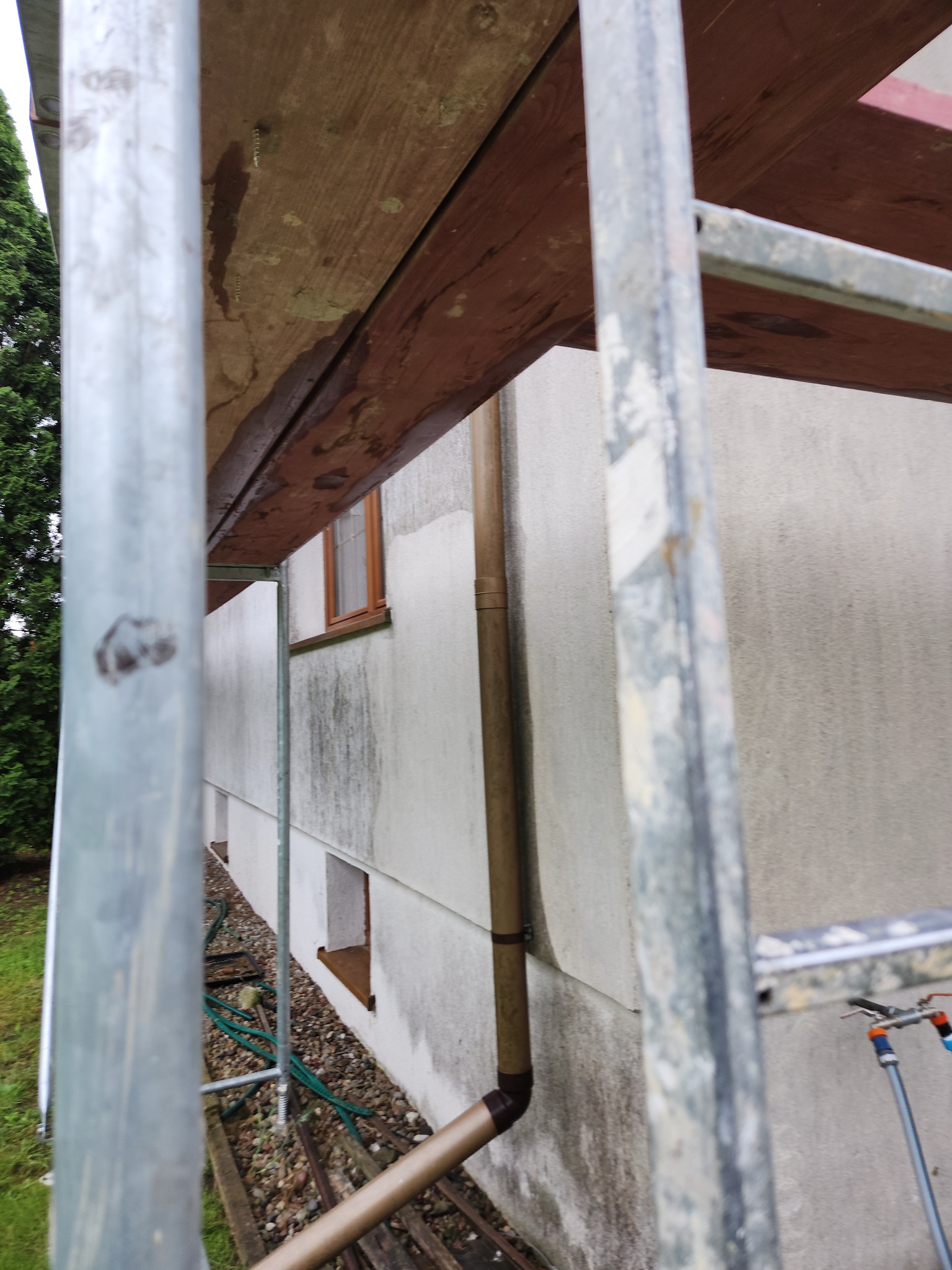 Elewacja domu w trakcie czyszczenia, widoczna z rusztowania. Brudna ściana z oknami i rurą spustową, elementy rusztowania na pierwszym planie.