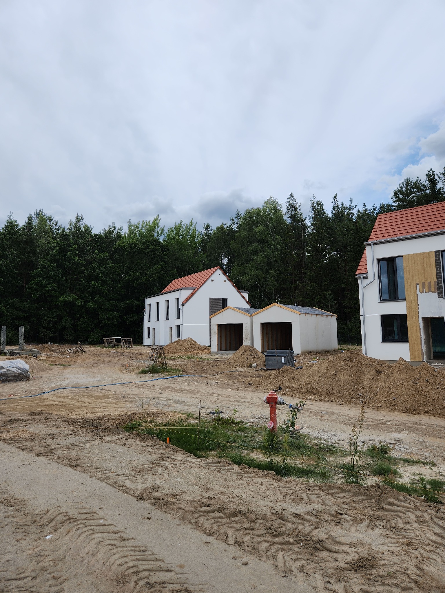 Dwa białe domy z czerwoną dachówką w trakcie budowy, widoczne z ziemi. Jeden z budynków ma częściowo wykończoną elewację. W tle las i pochmurne niebo.