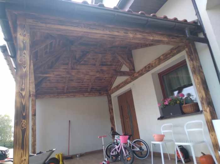 Zadaszenie tarasu z drewnianą konstrukcją i widoczną więźbą dachową, rynna wzdłuż krawędzi dachu. Na tarasie rower, hulajnoga i inne przedmioty.