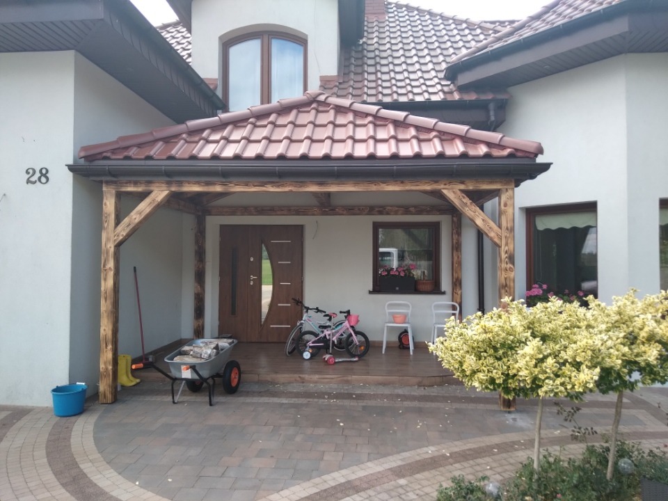 Zadaszenie nad wejściem do domu z brązową dachówką i drewnianą konstrukcją. Na podjeździe taczka z materiałami, rower dziecięcy i krzesła. Widoczne detale wykończenia dachu.