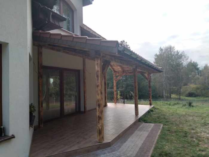 Zadaszony taras z drewnianymi słupami i fragmentem dachu z brązową dachówką, przylegający do jasnego budynku. Widok na zielony ogród.