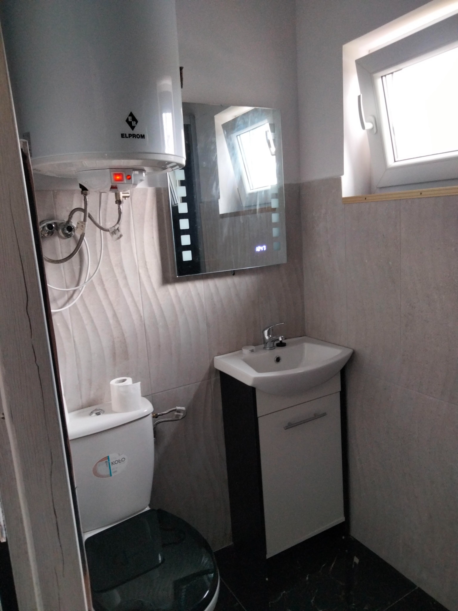 Nowoczesna łazienka z białą armaturą: wiszący bojler Elprom, umywalka z szafką i toaleta Koło na tle płytek imitujących kamień.