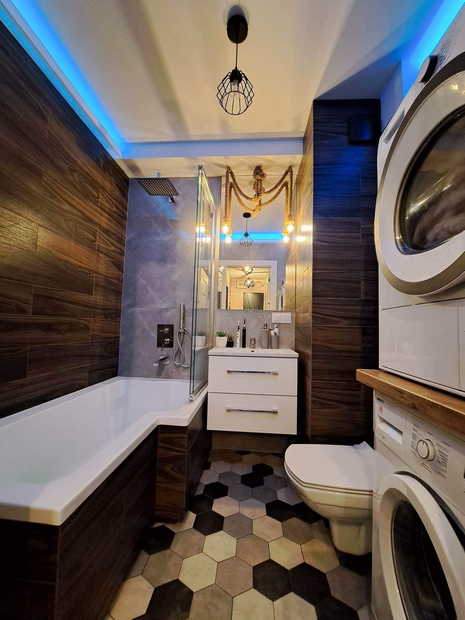 Nowoczesna łazienka z drewnopodobnymi płytkami, heksagonalną podłogą, podświetleniem LED i lampami z motywem liny, z pralką i suszarką w rogu.