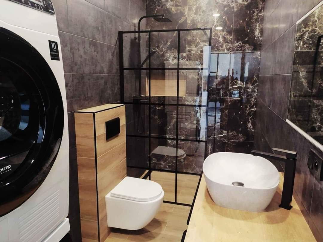Nowoczesna łazienka z marmurowymi płytkami, kabiną prysznicową z czarną ramą, umywalką nablatową, toaletą podwieszaną z drewnianą obudową i pralką.