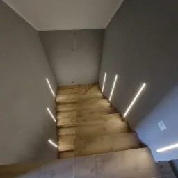 Klatka schodowa z drewnianymi schodami oświetlonymi liniowymi lampami LED wbudowanymi w ściany, szare ściany i biały sufit, nowoczesny design.