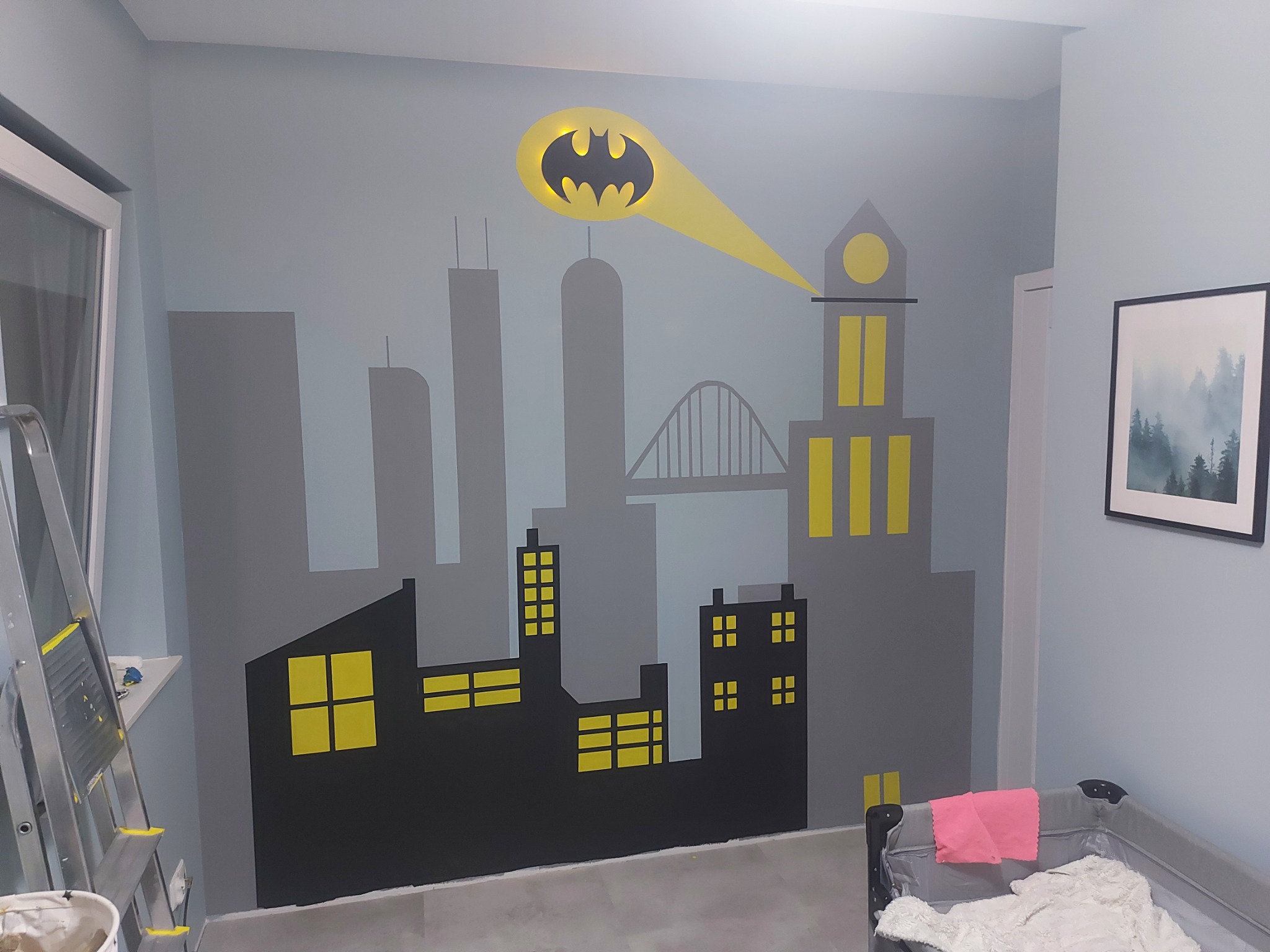 Ściana w pokoju dziecięcym w Wejherowie z ręcznie malowanym motywem Gotham City i symbolem Batmana, oświetlonym od tyłu, widoczna drabina malarska i łóżeczko dziecięce.
