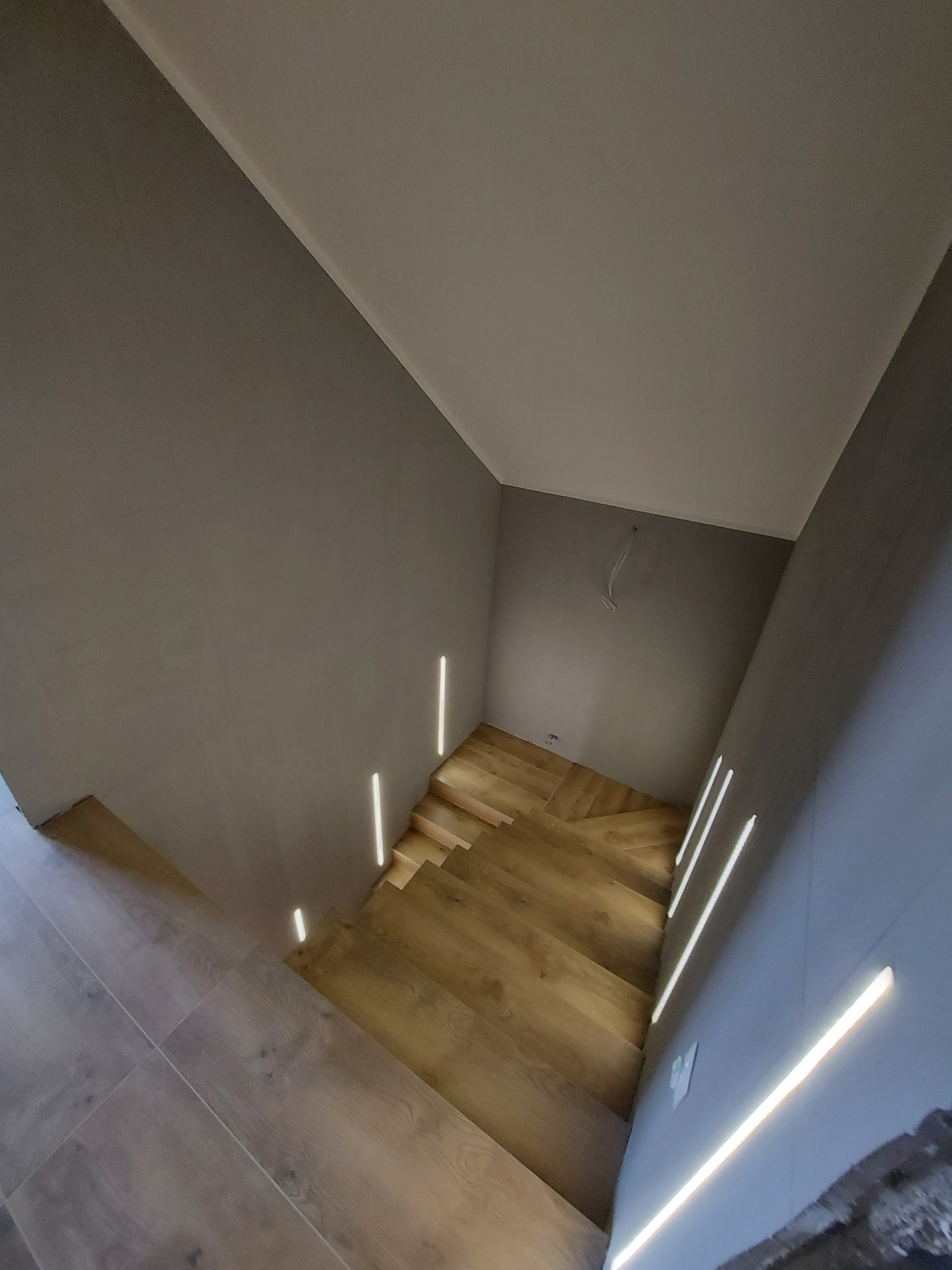 Nowoczesne schody z drewnianymi stopniami i podświetleniem LED wzdłuż ściany, szara ściana i jasny sufit, widok z góry.