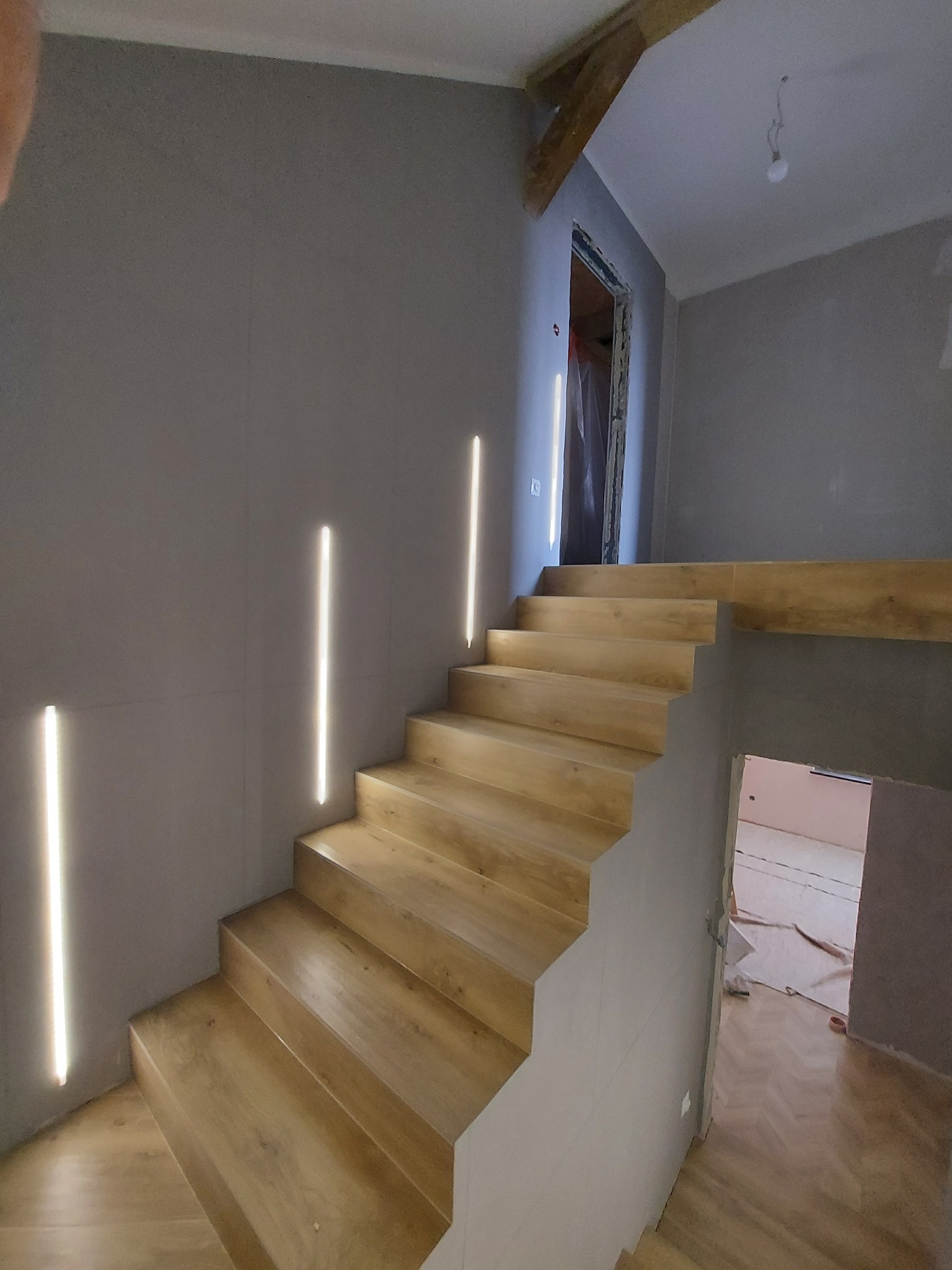 Drewniane schody z podświetleniem LED w ścianie, szare ściany, widoczne elementy konstrukcyjne sufitu, pomieszczenie w trakcie remontu.