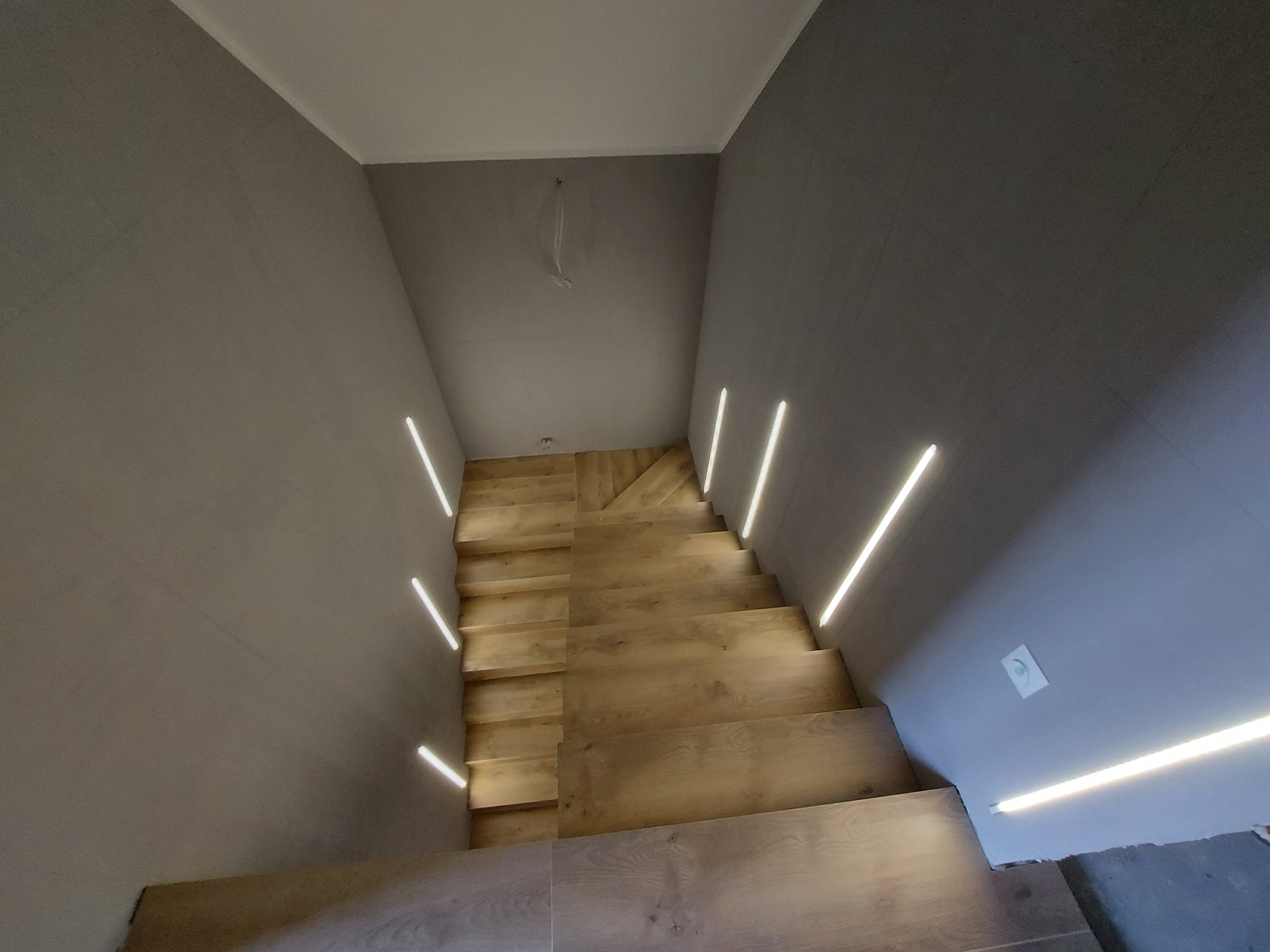 Klatka schodowa z drewnianymi schodami oświetlonymi liniowymi lampami LED wbudowanymi w ściany, szare ściany i biały sufit, nowoczesny design.