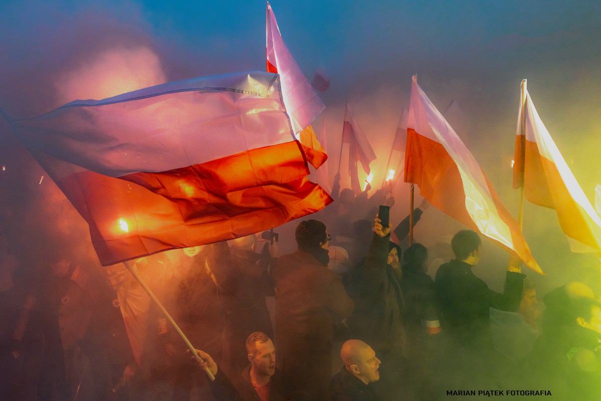 Tłum ludzi z polskimi flagami w dymie i z flarami, z widocznymi twarzami w dolnej części kadru.