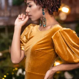 Portret kobiety w złotej, welurowej sukience z bufkami i wiszącymi kolczykami, zamyślona poza, miękkie światło i świąteczne tło z bokeh.