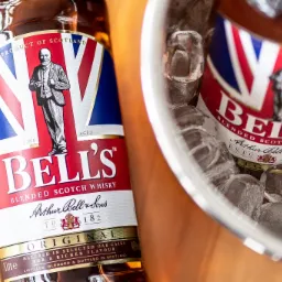 Dwie butelki szkockiej whisky Bell's: jedna na drewnianym stole, druga chłodzona w srebrnym wiaderku wypełnionym lodem.