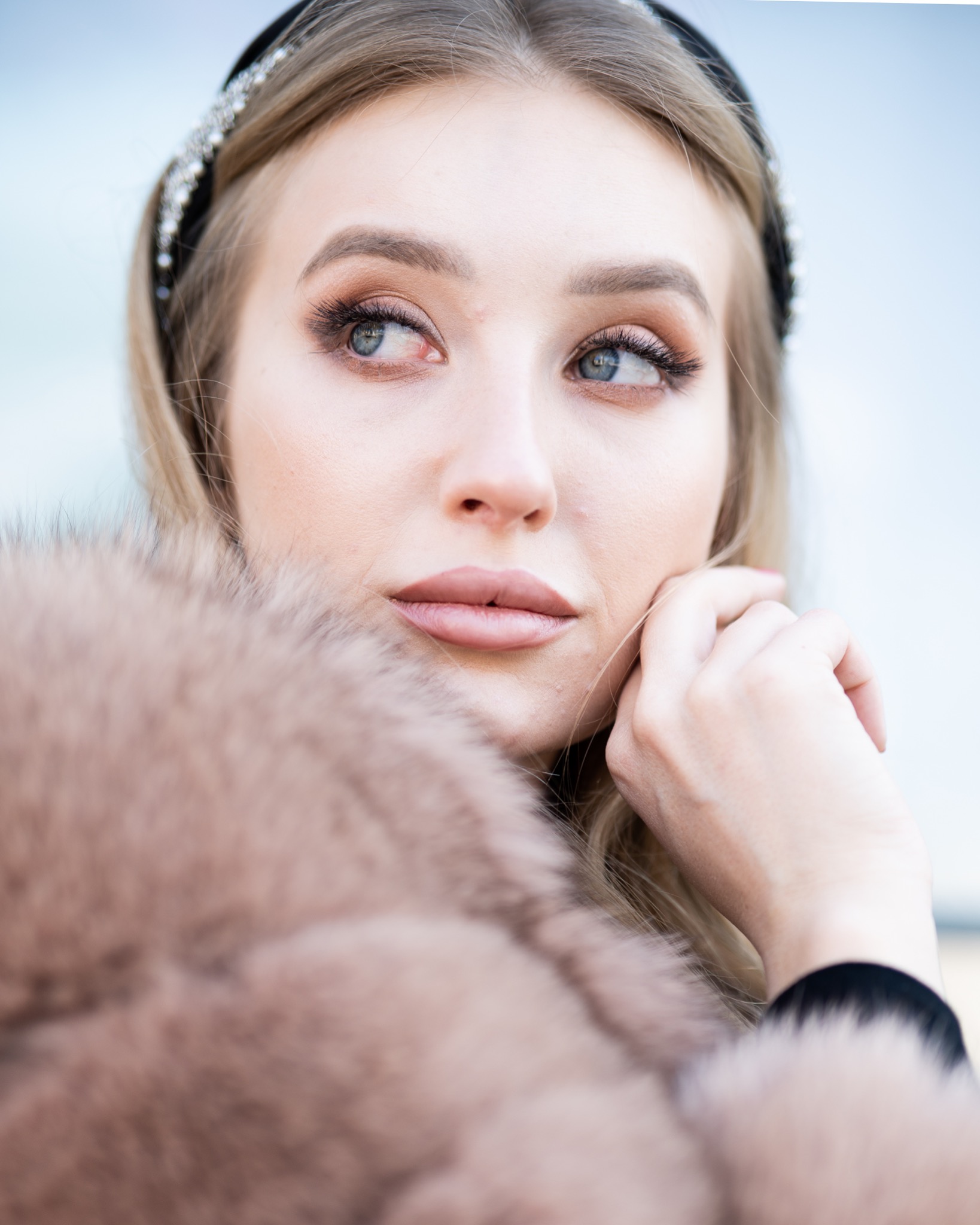 Portret kobiety w futrze z delikatnym makijażem i ozdobną opaską, zamyślona poza i subtelne oświetlenie.