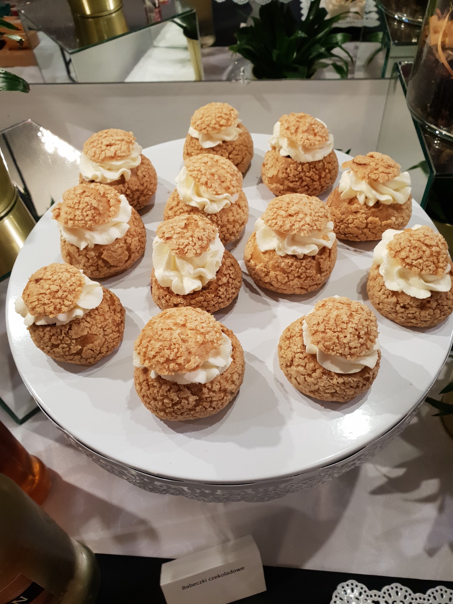 Kremowe ptysie na białym, ozdobnym stojaku, widoczne z góry, z delikatnym tłem roślinnym i elementami dekoracyjnymi.