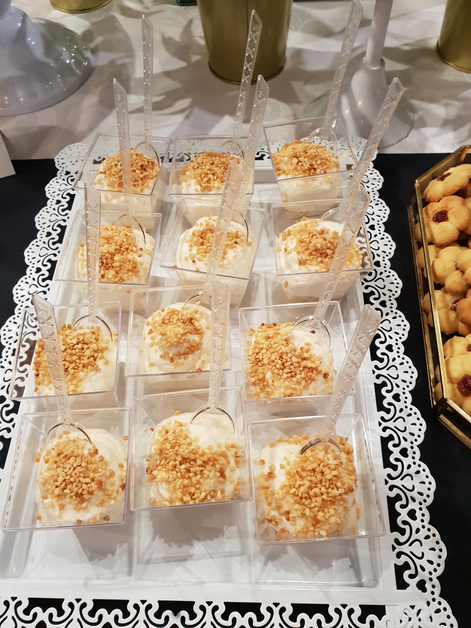 Deser w małych, przezroczystych pojemnikach, posypany orzechami, z plastikową łyżeczką, ułożony na białej, ażurowej tacy, obok ciastek z dżemem, oferowany w Sztumie.