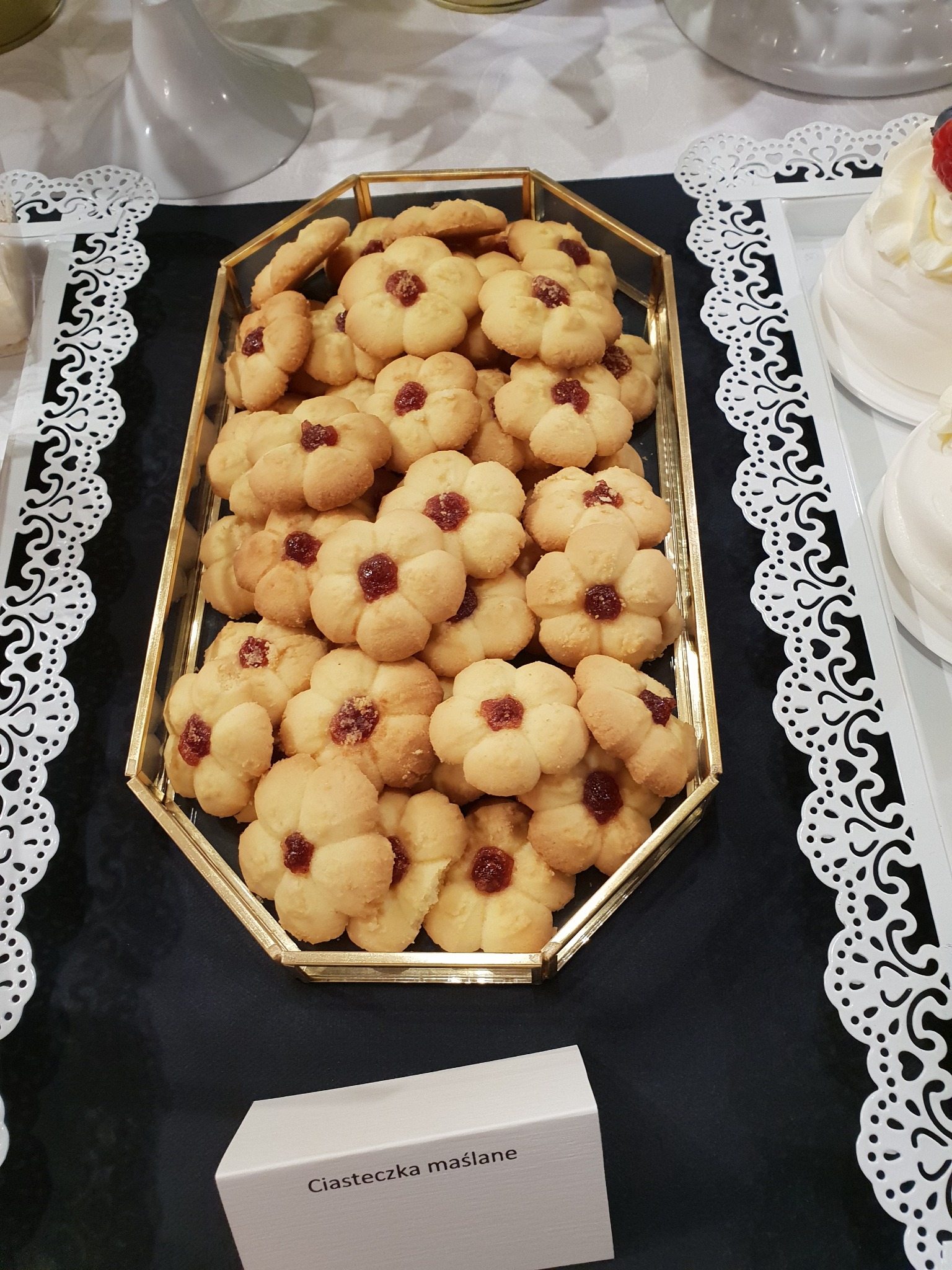 Ciastka maślane w kształcie kwiatów z marmoladą w złotym, geometrycznym naczyniu, obok deser bezowy na białej tacy z koronkową obwódką, na czarnym obrusie.