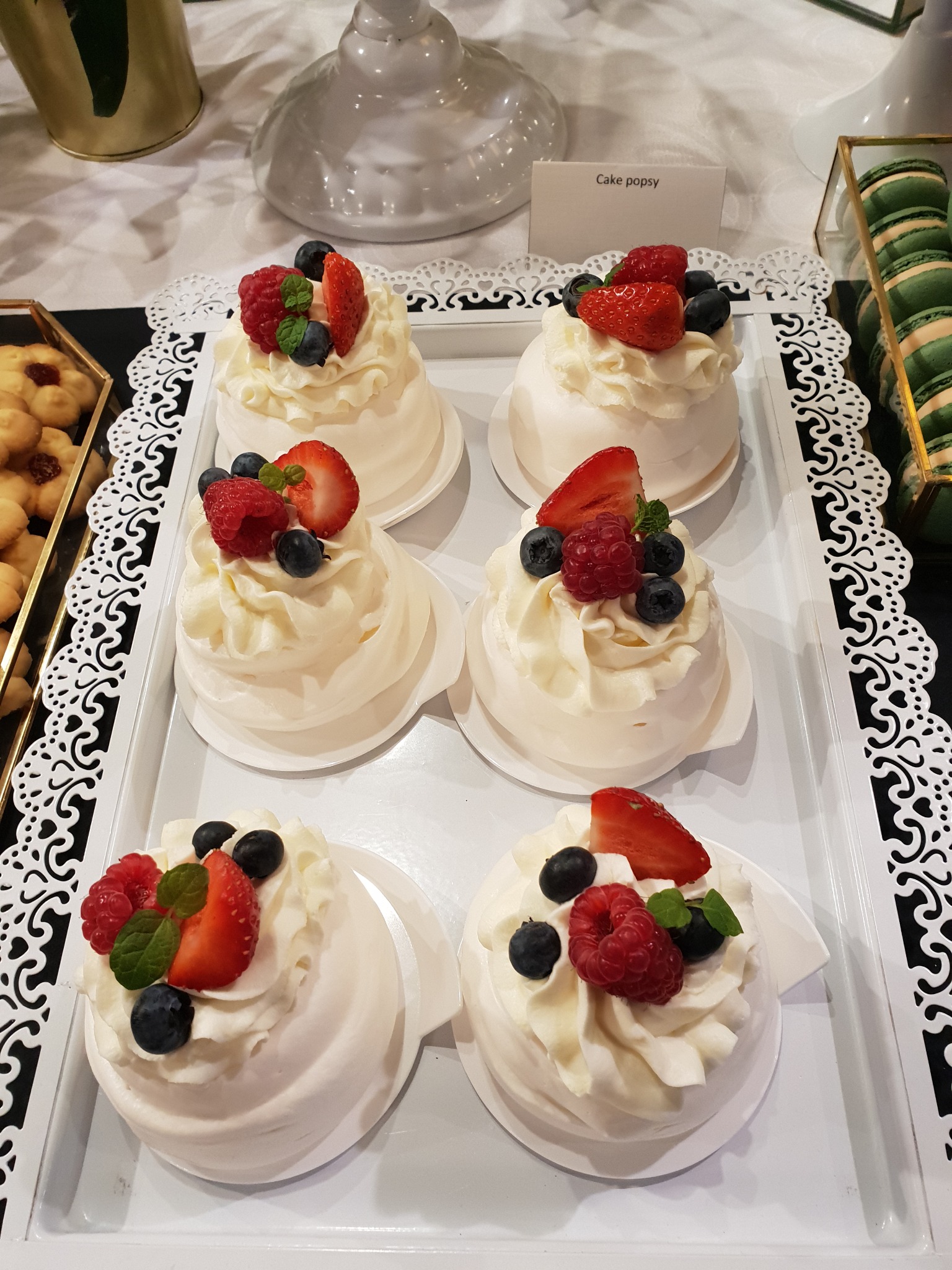 Sześć mini bezowych tortów Pavlova na białej tacy, udekorowanych bitą śmietaną, truskawkami, malinami i borówkami, z dekoracyjną białą serwetką i pudełkami z ciasteczkami i makaronikami w tle.